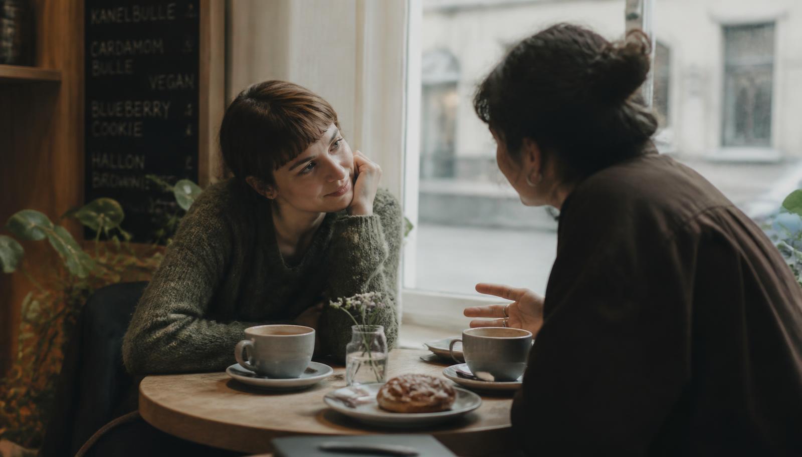 Two people leaning in across a small round café table, one listening with genuine attention, coffee cups and a cinnamon bun between them in soft daylight.